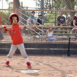 kid in red uniform swinging a baseball bat about to hit a ball